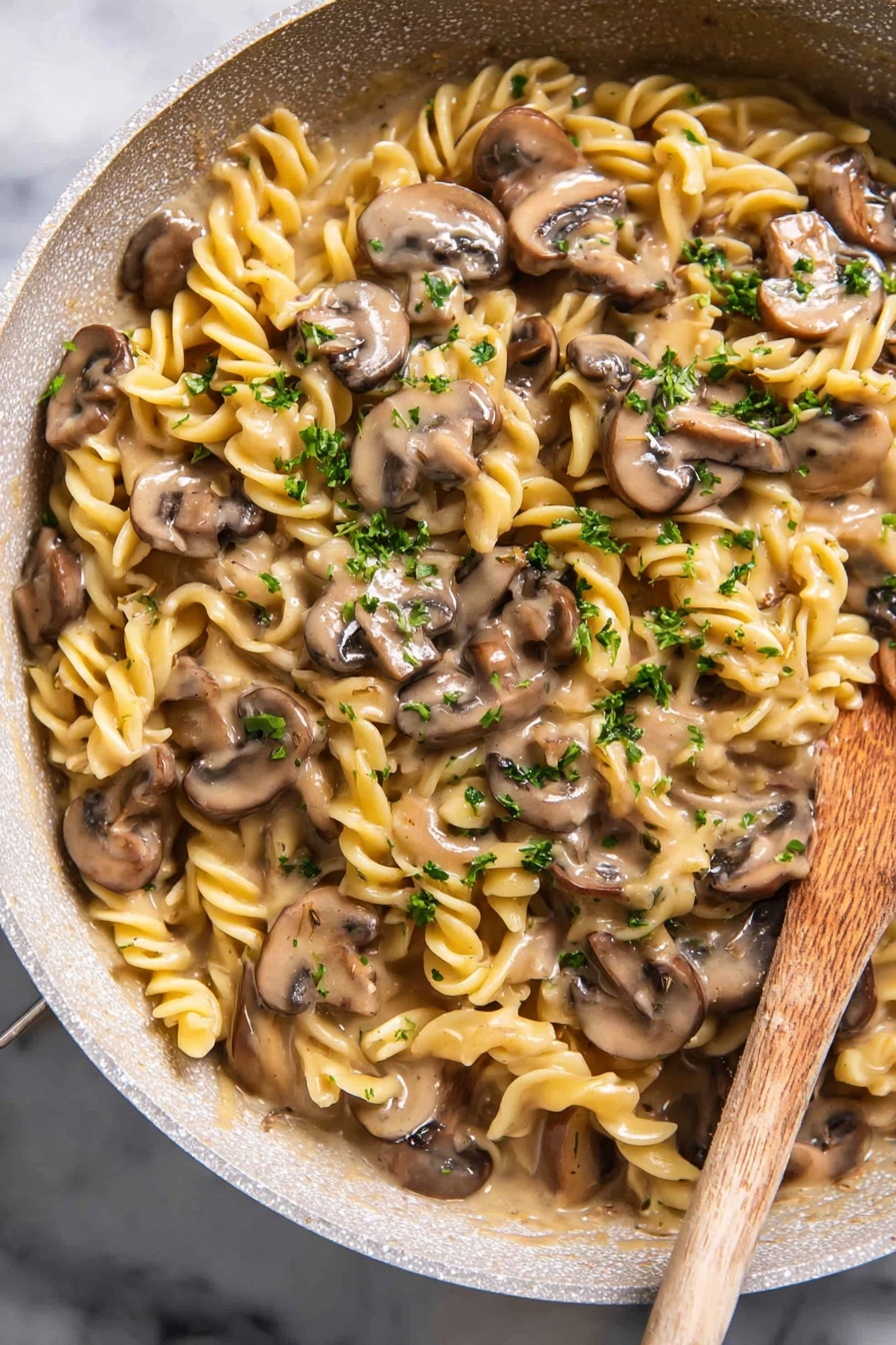 Vegan Mushroom Stroganoff - The image shows a close-up of a white pot filled with creamy mushroom pasta. The pasta is spiral-shaped and light yellow, mixed evenly with sliced brown mushrooms in a thick, beige cream sauce. Small green parsley bits are scattered on top, adding color contrast. A wooden spoon is partially visible, resting inside the pot, coated with some sauce and pasta. The pot is placed on a white marbled surface, with two whole mushrooms and some parsley leaves blurred in the background. Photo taken with an iphone --ar 2:3 --v 7