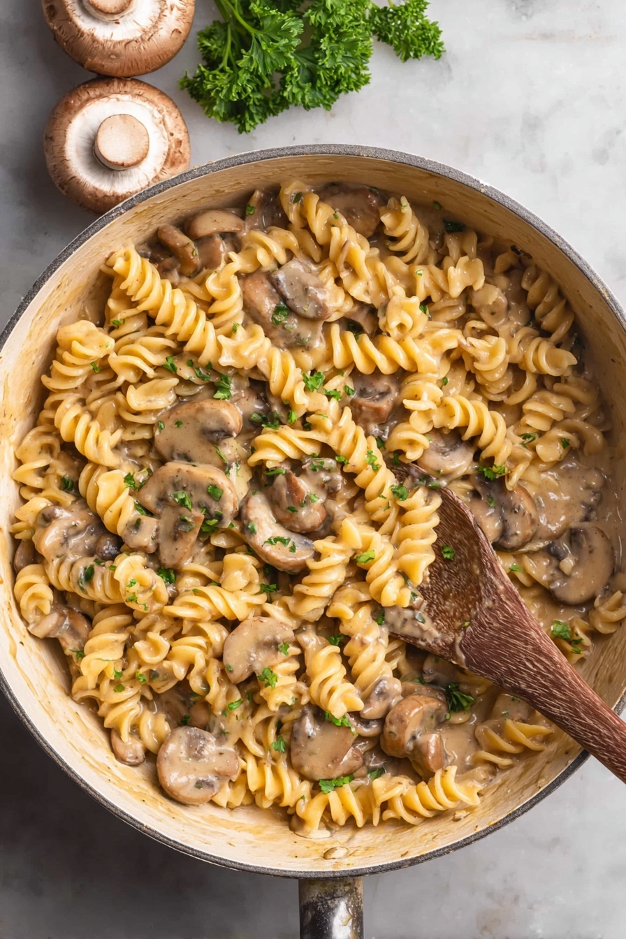 Vegan Mushroom Stroganoff - The image shows a close-up of a pan filled with creamy pasta and mushrooms. The pasta is light yellow and twisted, spreading evenly across the pan. Sliced brown mushrooms with soft, smooth surfaces are mixed with the pasta. A light brown creamy sauce coats everything, making the dish look rich and smooth. Small bits of green parsley are sprinkled over the top, adding a fresh touch of color. A wooden spoon is partly visible on the right side, resting inside the pan. The pan is white with a subtle textured finish, and the surface below is a white marbled texture. Photo taken with an iphone --ar 2:3 --v 7
