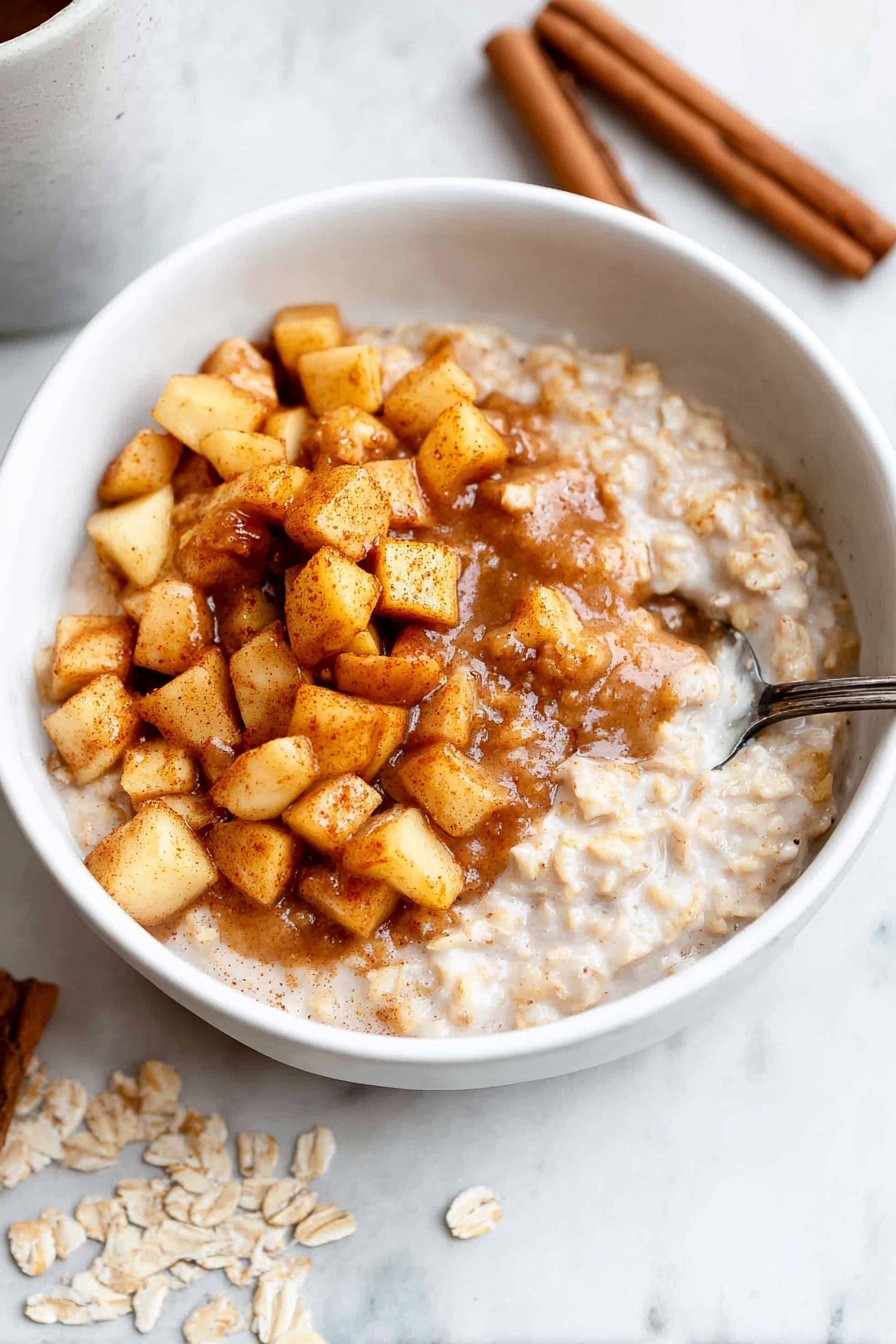 A white bowl filled with creamy oatmeal forms the base layer, with a thick, smooth texture and light beige color. On top, there is a generous heap of small, golden-brown cooked apple cubes mixed with a rich, warm cinnamon sauce that spreads slightly into the oatmeal beneath. A silver spoon is scooping some of the layered oatmeal and apples from the right side of the bowl. The bowl is placed on a white marbled surface; near it lie three cinnamon sticks and some scattered rolled oats. Photo taken with an iphone --ar 2:3 --v 7