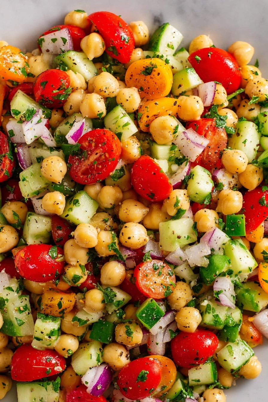 A white bowl filled with a colorful salad showing about five layers of ingredients. The first layer has light beige chickpeas scattered all over. The second layer has bright red grape tomatoes, cut in half, mixed throughout. The third layer contains light green chunks of cucumber, cut irregularly. The fourth layer includes dark green diced bell peppers. The fifth layer has small pieces of purple-red onion spread evenly. Everything is sprinkled with finely chopped green parsley and black pepper. The bowl rests on a white marbled surface with part of a lemon and some parsley visible in the background, along with a gold spoon inside the bowl. Photo taken with an iphone --ar 2:3 --v 7