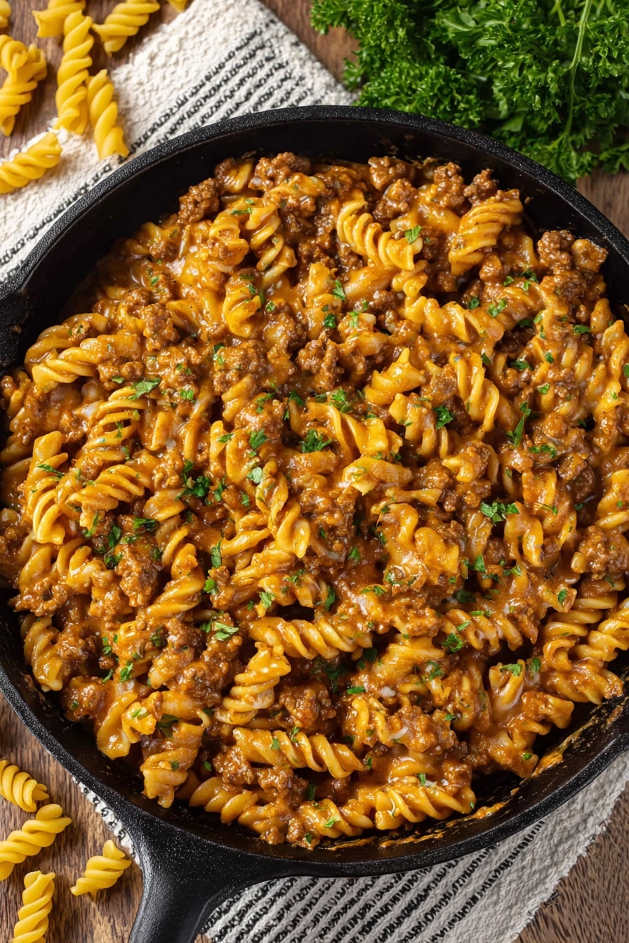 Vegan Cheeseburger Pasta - The image shows a black cast iron pan filled with a creamy pasta dish made of spiral pasta mixed with browned ground meat in a rich, orange-tan sauce. Small pearl-like pieces give texture throughout the dish, and green parsley leaves are scattered on top for color. The pan is placed on a textured white and black striped cloth on a wooden surface, with some uncooked spiral pasta and fresh parsley visible nearby. The colors are warm and inviting, and the texture of the sauce looks smooth and thick, coating the pasta evenly. photo taken with an iphone --ar 2:3 --v 7