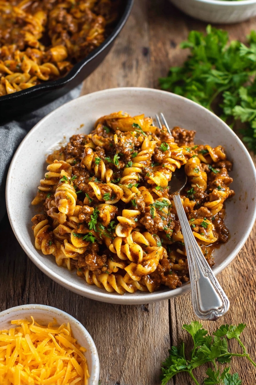 Vegan Cheeseburger Pasta - A black cast iron skillet filled with a creamy pasta dish that has three visible layers: the bottom layer shows spiral pasta in a light yellow color, the middle layer is a rich, brown plant-based meat sauce mixed evenly with the pasta, and the top layer is melted cheese in golden orange with small green herb sprinkles scattered on top. The skillet is placed on a black and white striped cloth over a white marbled surface. Around the skillet, uncooked spiral pasta pieces are scattered, and a small white bowl filled with shredded orange cheese is nearby. A tray of raw plant-based ground meat with a pinkish tone is also partially visible in the top left corner of the image. Photo taken with an iphone --ar 2:3 --v 7