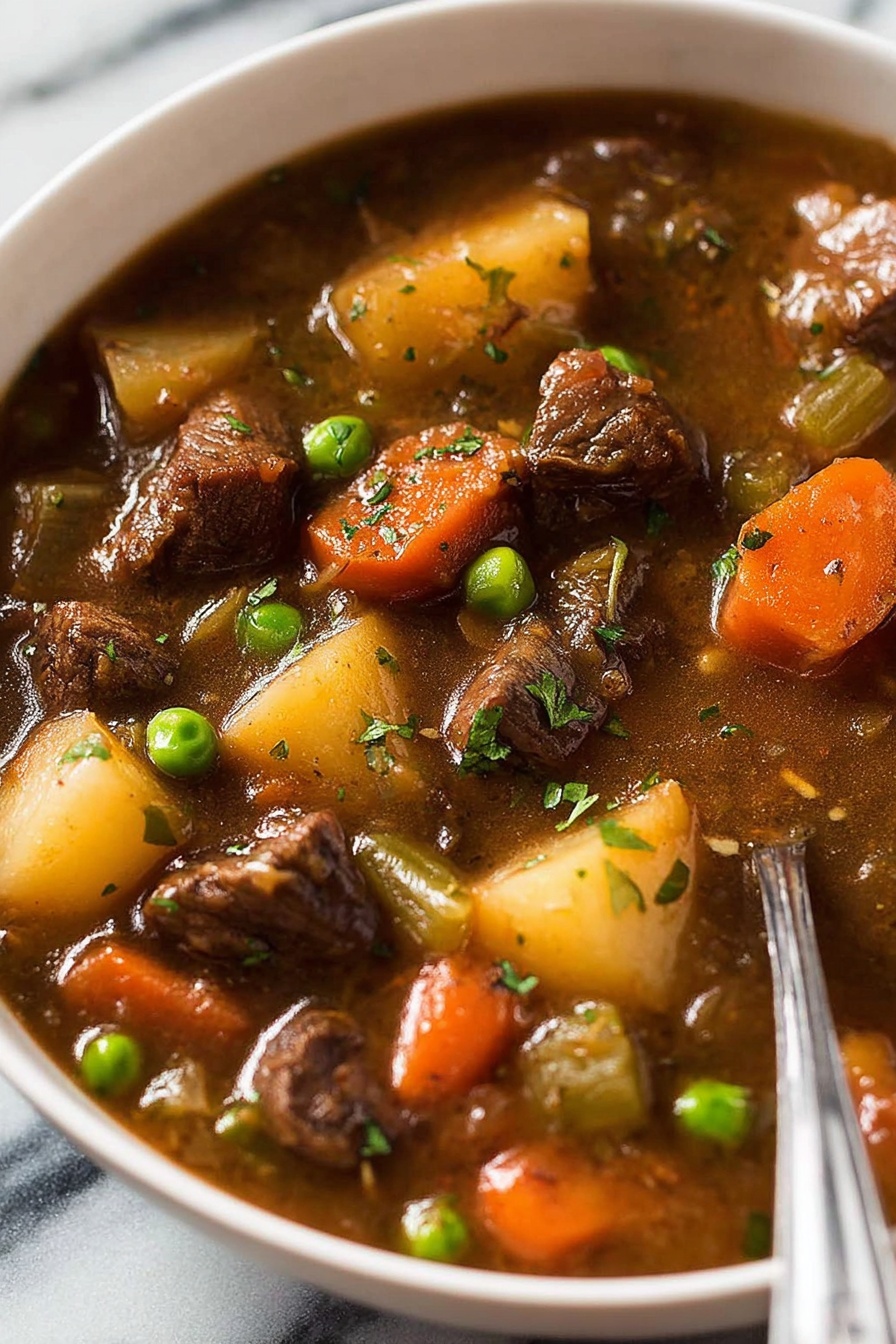 The image shows a close-up of a bowl filled with beef stew. The dish has chunks of dark brown beef, bright orange carrot pieces, light yellow potato cubes, green peas, and small pieces of celery in a thick brown broth. The ingredients look cooked and tender, with some chopped green herbs sprinkled on top. The bowl is white and placed on a white marbled surface, and a metal spoon is partially visible on the right side. photo taken with an iphone --ar 2:3 --v 7