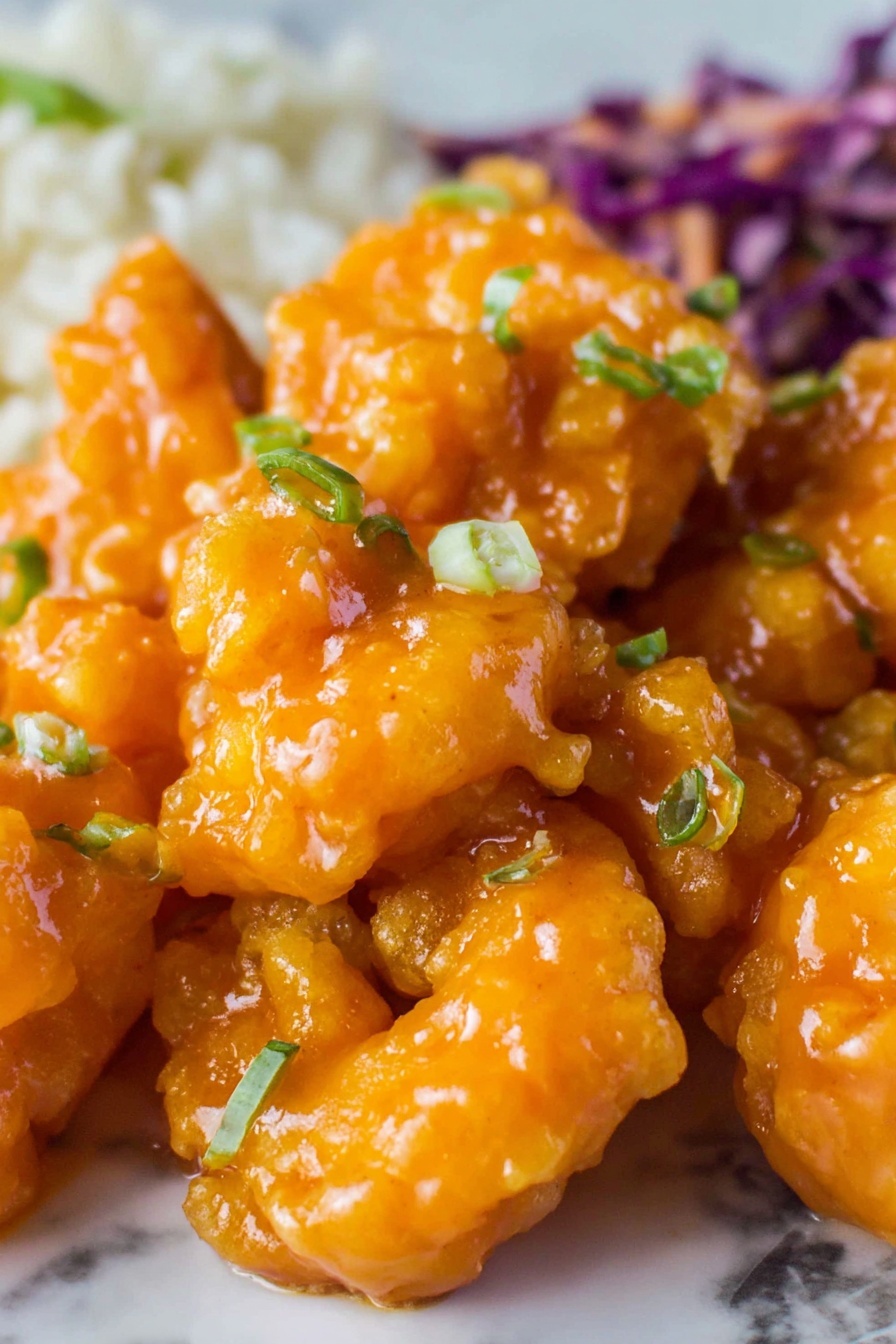 A close-up of a single orange-colored shrimp coated in a shiny, thick sauce being held by light brown wooden chopsticks. The shrimp shows a bumpy texture from the sauce with tiny green herb bits on it. The background is filled with more shrimp in the same sauce, slightly blurred. All is set against a white marbled surface. photo taken with an iphone --ar 2:3 --v 7