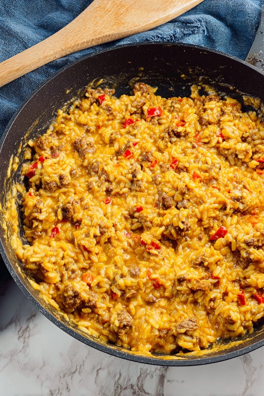 Inside a black pan, there is a creamy, thick dish made of rice mixed with small chunks of browned meat and bits of red pepper scattered throughout. The rice is coated in a yellow-orange sauce that looks rich and smooth, with some pieces of melted cheese visible at the bottom edge, blending into the other ingredients. The pan rests on a white marbled surface with a blue cloth and a wooden spatula nearby. Photo taken with an iphone --ar 2:3 --v 7