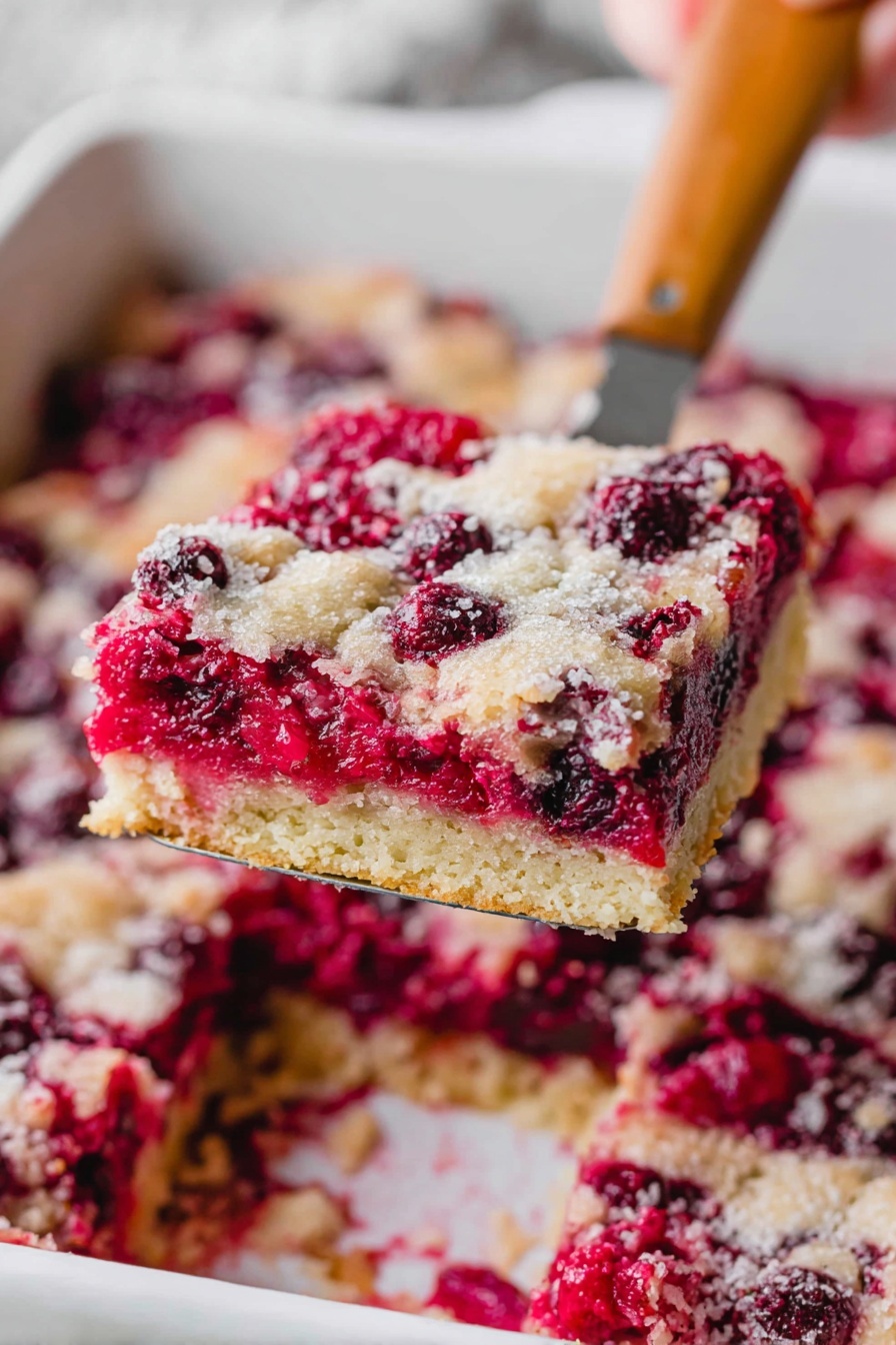 A close-up of a square piece of berry dessert being lifted by a spatula above a white baking dish. The dessert has three layers: a light golden, crumbly bottom crust; a middle layer of bright red and purple whole berries that look juicy and soft; and a top layer that is lightly browned and textured with small patches of baked batter and scattered berries. The white marbled surface is blurred in the background, and a woman's hand is holding the light wooden-handled spatula. Photo taken with an iphone --ar 2:3 --v 7