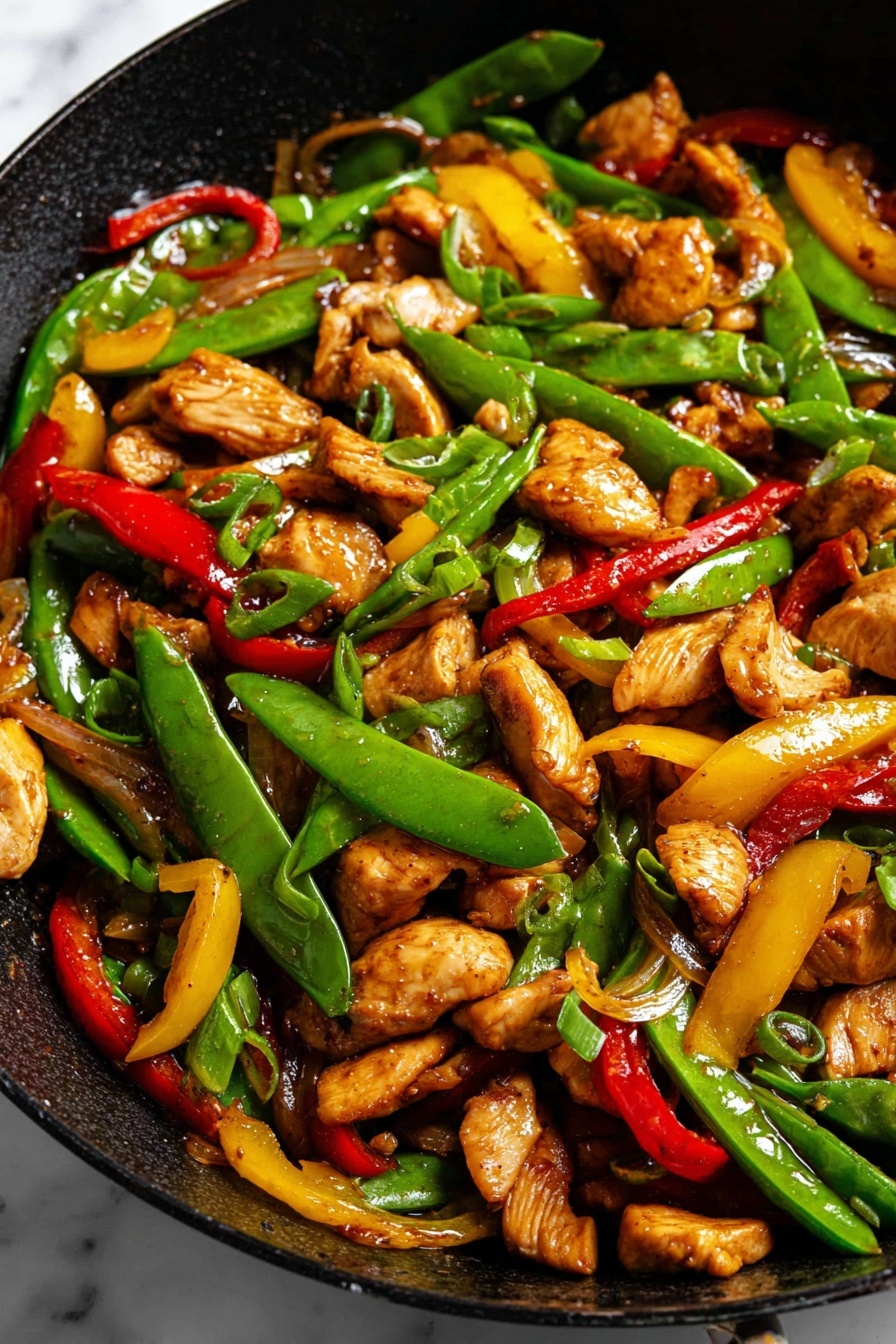 This is a close-up of a black pan filled with stir-fried pieces of cooked light brown chicken mixed with bright green snap peas, thin slices of red and yellow bell peppers, and green onion pieces. The chicken pieces have a slightly shiny, cooked texture and are mixed throughout the vibrant green vegetables. The pan is set on a white marbled surface. photo taken with an iphone --ar 2:3 --v 7