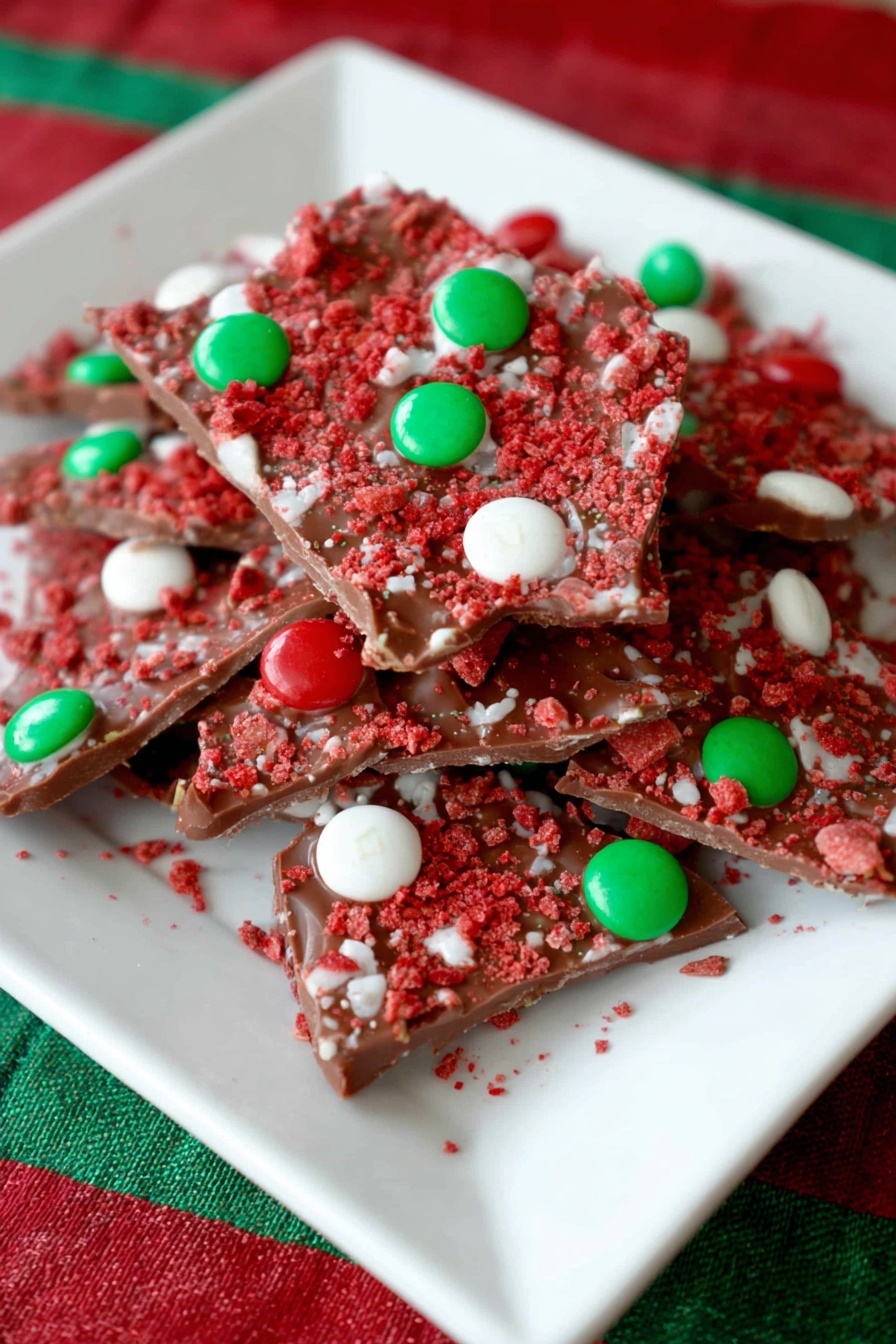 A white square plate holds a pile of irregular broken pieces of milk chocolate bark. The bark is topped with a layer of red crushed crumbs that add a rough texture across all pieces. Scattered on top are small round candies in red, green, and white colors, embedded into the chocolate surface. The background shows a white marbled texture, and the plate rests on red and green striped fabric. photo taken with an iphone --ar 2:3 --v 7