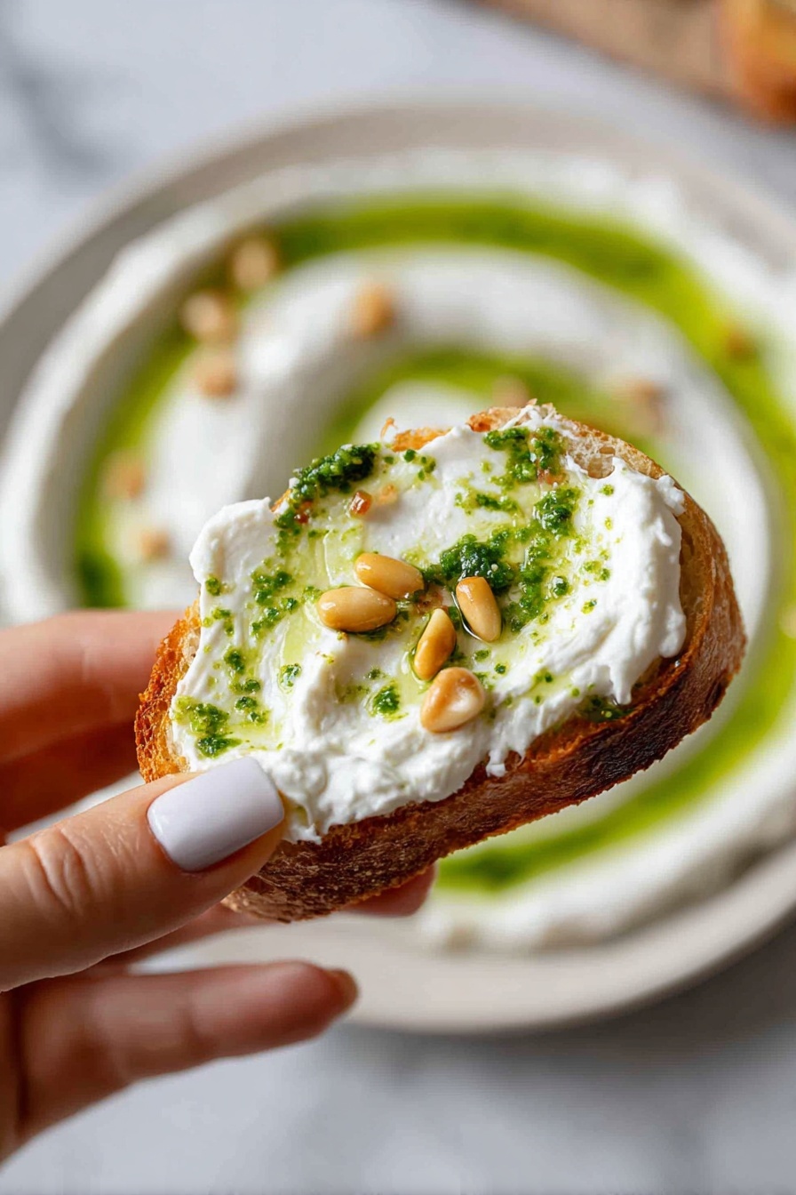 A woman's hand holding a toasted bread slice topped with a thick layer of white creamy spread, drizzled with bright green herb oil and a single golden pine nut; in the blurred background, a white plate with concentric circles of the same creamy white spread and green oil, sprinkled with small golden pine nuts, all on a white marbled surface. photo taken with an iphone --ar 2:3 --v 7