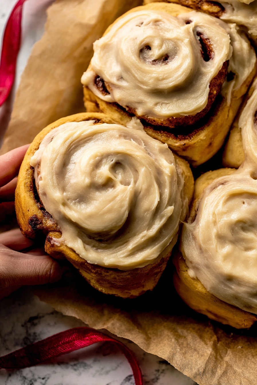 The image shows a close-up of four cinnamon rolls arranged tightly together on a piece of light brown parchment paper, placed on a white marbled surface. Each cinnamon roll has a dark golden-brown dough base with visible cinnamon swirls inside. On top, there is a thick, smooth layer of light brown frosting with a creamy texture that softly covers the rolls, showing slight swirling patterns from spreading. A woman's hand is gently holding the bottom left cinnamon roll, adding a human touch to the cozy scene. A thin red ribbon lies partially under the parchment paper. photo taken with an iphone --ar 2:3 --v 7