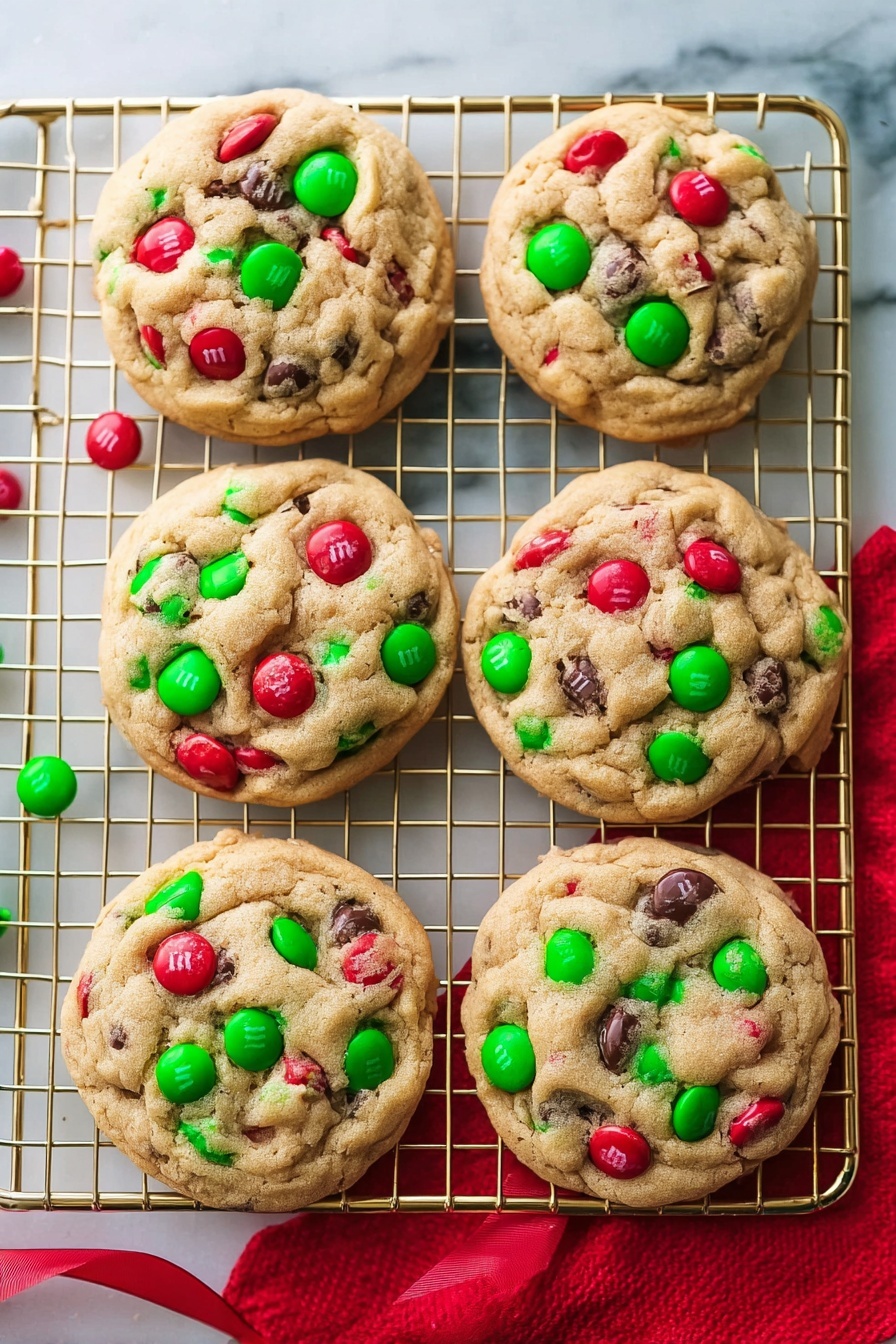 Six round cookies are placed on a gold cooling rack over a white marbled surface. Each cookie has a light brown dough base with red and green candy-coated chocolates scattered on top and partially embedded, making the surface colorful and textured. Some candies have a small white