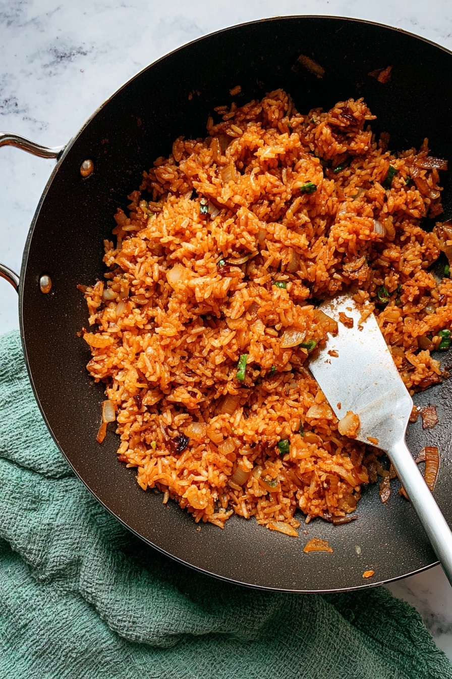 A black wok filled with a pile of orange-red fried rice mixed with small pieces of light brown onions and green herbs, showing a moist and slightly shiny texture, with a metal spatula resting inside on the right side. The wok sits on a soft, textured green cloth. The background is a white marbled surface. photo taken with an iphone --ar 2:3 --v 7