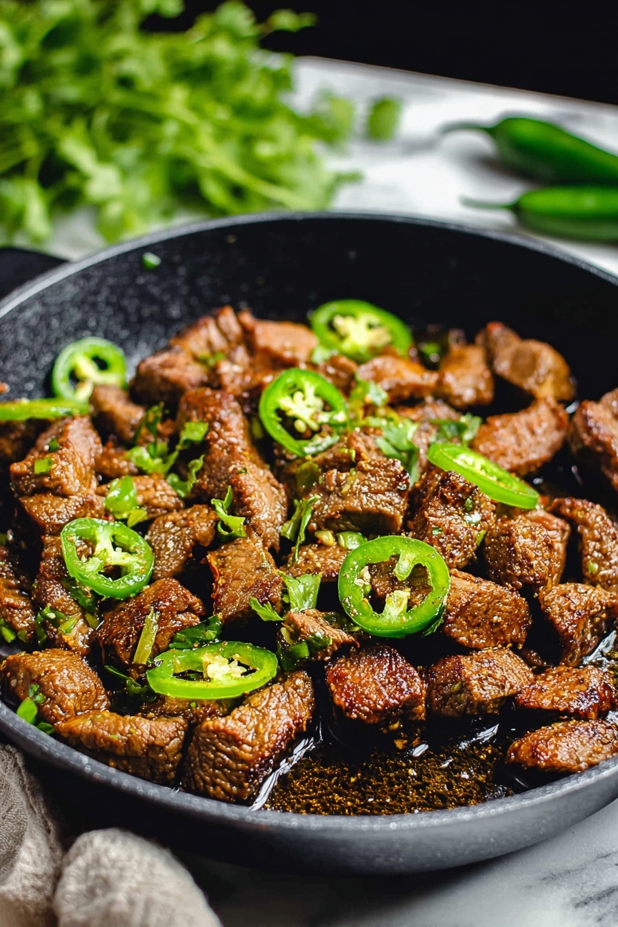 The image shows a black pan filled with browned, small pieces of cooked meat, which are unevenly shaped and textured with a slightly crispy outside. Scattered on top and mixed among the meat pieces are bright green slices of jalapeño peppers, some whole rings and some in curved strips. There is a bit of oil or sauce pooling at the bottom of the pan, giving a shiny look to the meat. The background is dark with a blurred bunch of fresh green herbs on the side. The whole scene is set on a white marbled surface. Photo taken with an iphone --ar 2:3 --v 7