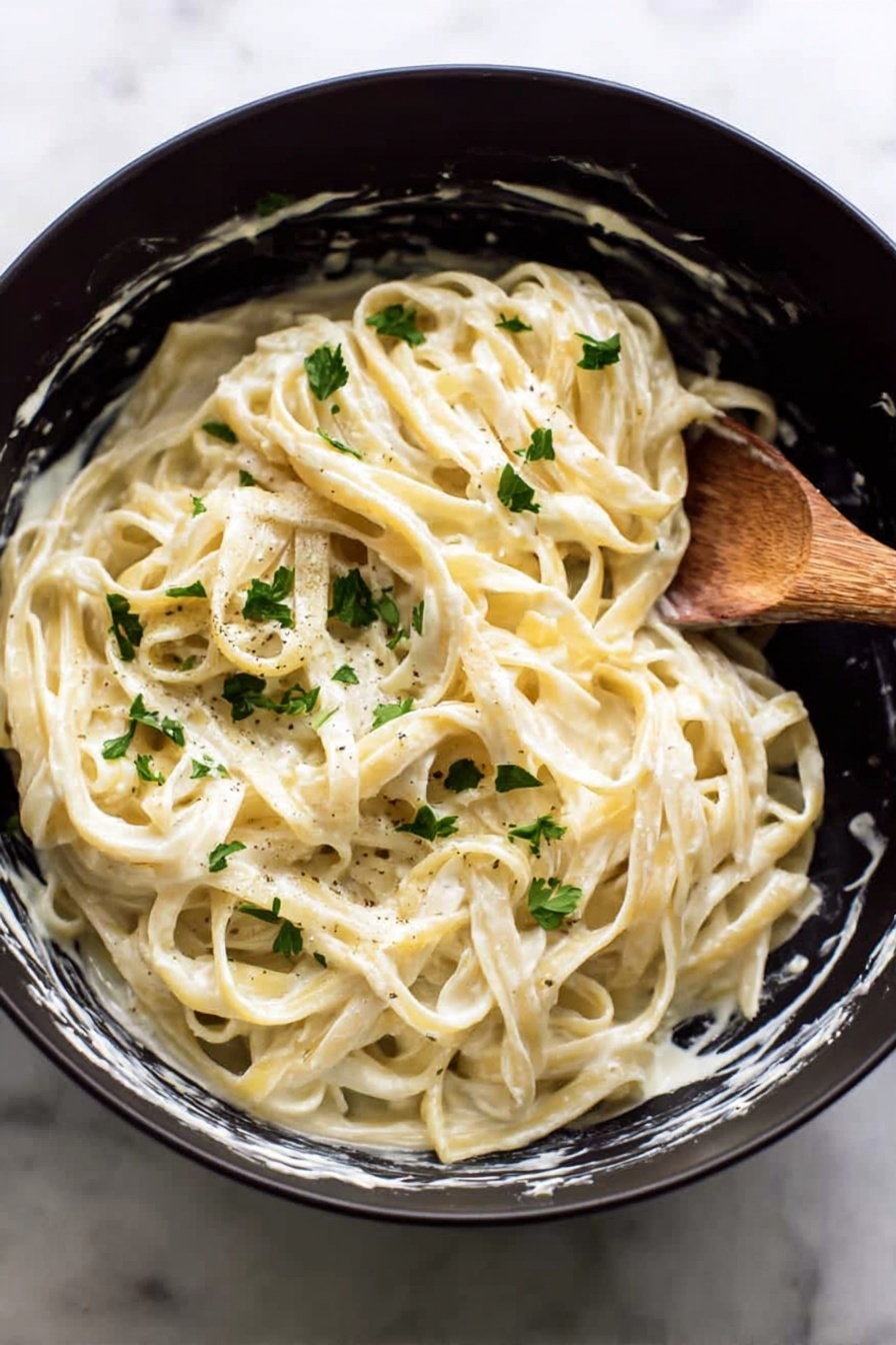 The image shows a black bowl filled with creamy fettuccine pasta mixed with white sauce, giving the noodles a smooth, shiny texture. There are small green parsley leaves sprinkled on top, adding a fresh bit of color. The edges of the bowl have some creamy sauce marks, suggesting the pasta has been freshly stirred. A wooden spoon is placed inside the bowl on the right side, partially covered with sauce. The bowl is set on a white marbled surface. Photo taken with an iphone --ar 2:3 --v 7