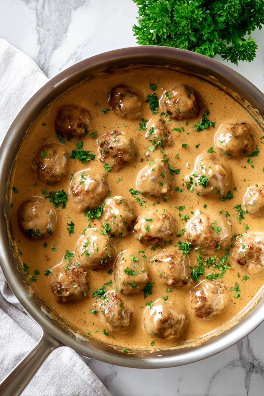 A silver pot filled with about three layers of round meatballs covered in thick, light brown gravy with a smooth texture. The meatballs have a browned, slightly rough surface and are partially submerged in the creamy sauce. Small green parsley leaves are sprinkled evenly on top, adding a fresh touch of color. The pot is placed on a white marbled surface with a sprig of fresh parsley nearby. Photo taken with an iphone --ar 2:3 --v 7