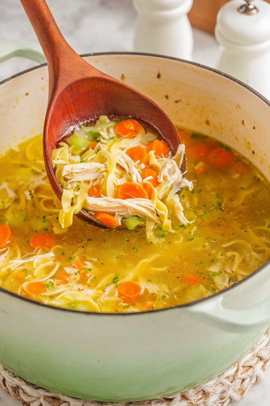 A close-up of a large pot filled with clear chicken soup that has a yellowish broth. The soup has layers of sliced orange carrots, light green celery pieces, shredded white chicken, and spiral-shaped yellow noodles, all resting on a wooden spoon lifted above the pot. The pot is white enameled with a soft green outer edge, sitting on a white marbled surface with a hint of white and beige woven mat underneath. The background is blurred, showing a white salt shaker and a pepper mill. photo taken with an iphone --ar 2:3 --v 7