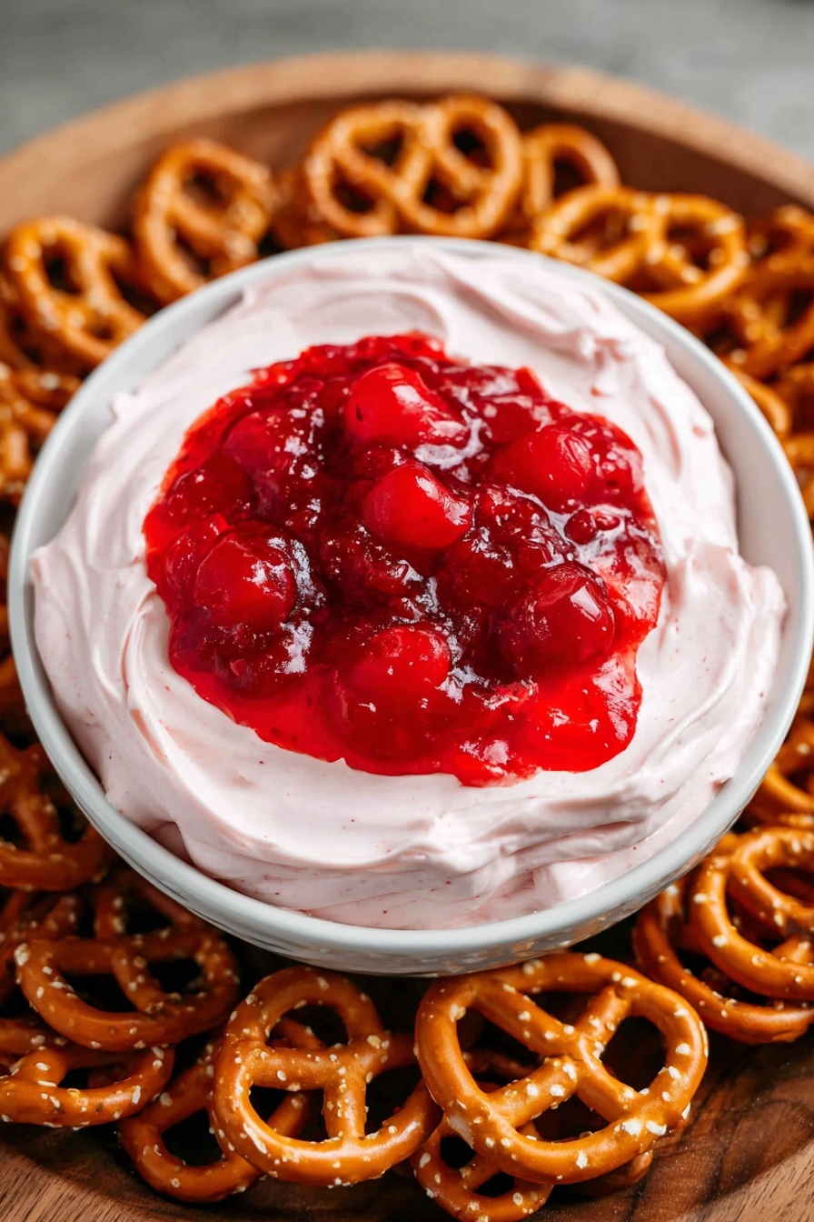 A white bowl with two layers sits in the center of a wooden tray filled with small golden brown pretzels. The first layer in the bowl is a smooth, light pink creamy dip that looks thick and soft, filling most of the bowl. On top of this dip is a glossy bright red cherry topping with whole cherries visible, creating a shiny, slightly wet texture. The wooden tray underneath is light brown and has a wood texture background. photo taken with an iphone --ar 2:3 --v 7